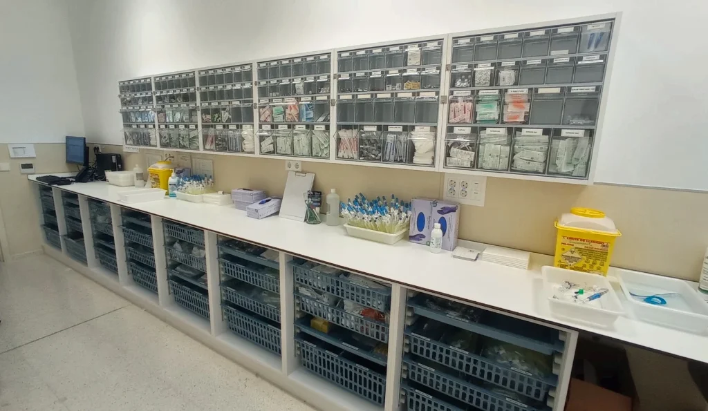 Sala de curas de un hospital con encimera corrida blanca, módulos bajos con cestas y cajetines de material en la pared