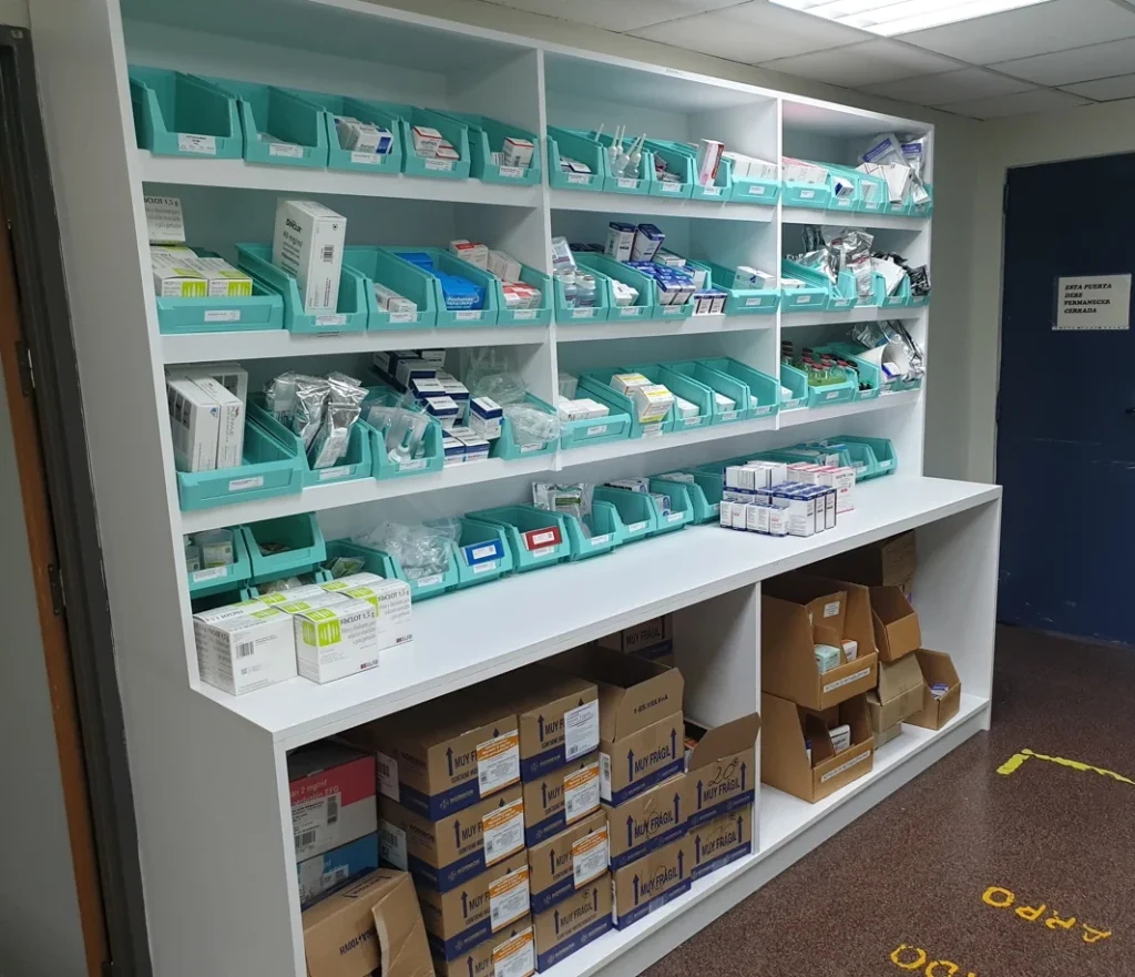 Almacén de farmacia hospitalaria con estanterías blancas, cubetas de medicación y encimera de trabajo sobre huecos abiertos
