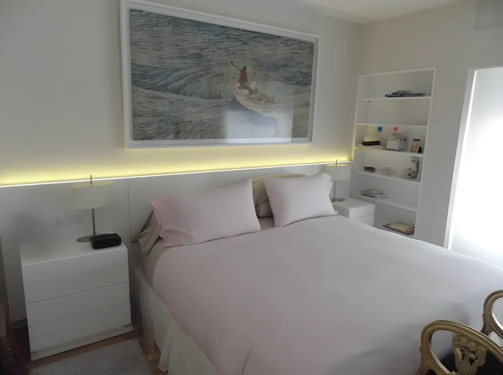 Bedroom with a white lacquered headboard with indirect LED lighting, bedside drawer units and a built-in bookcase